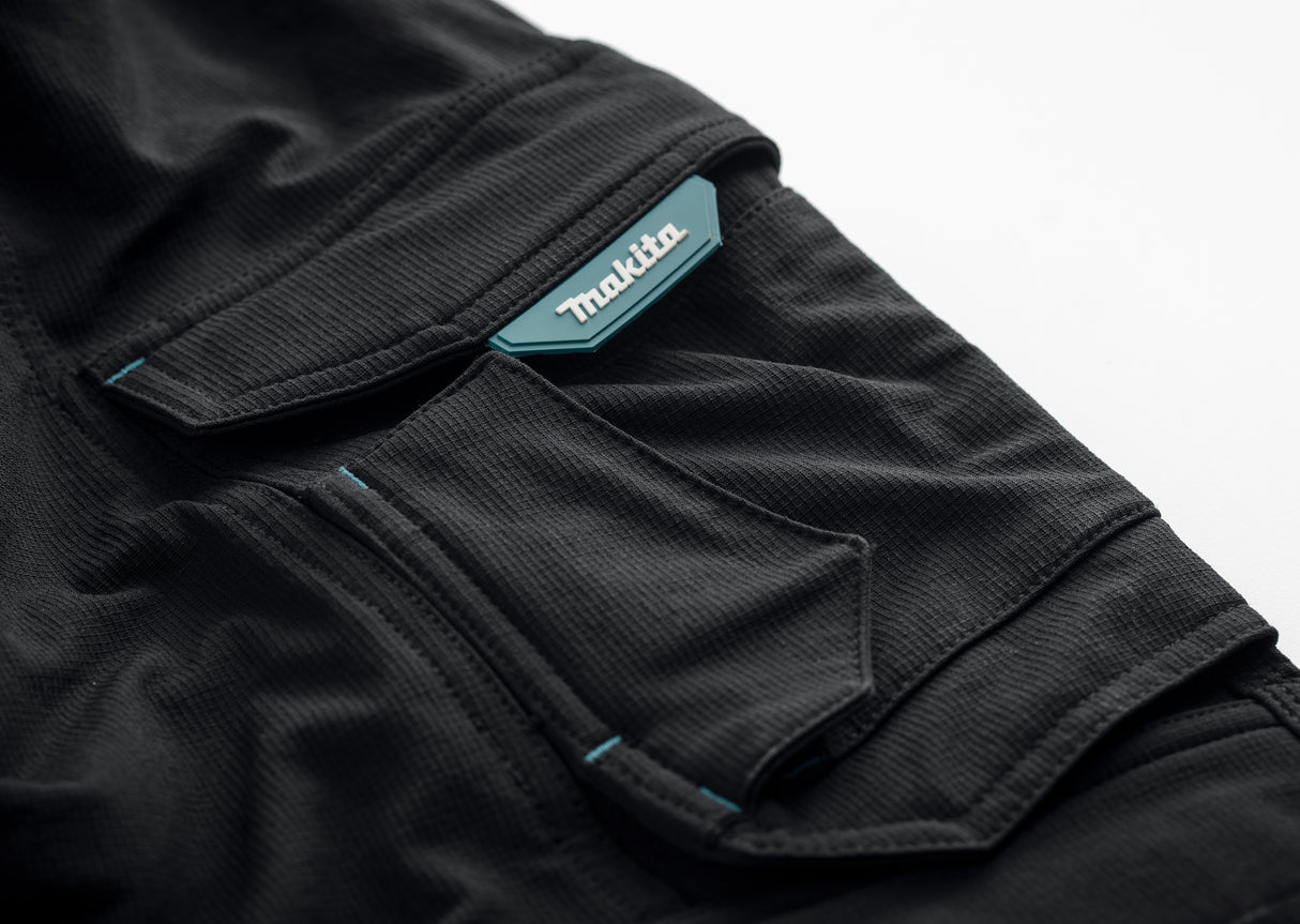 Close up of the pocket on the black Makita Onyx 4 way stretch trousers with a turquoise Makita label on a white background