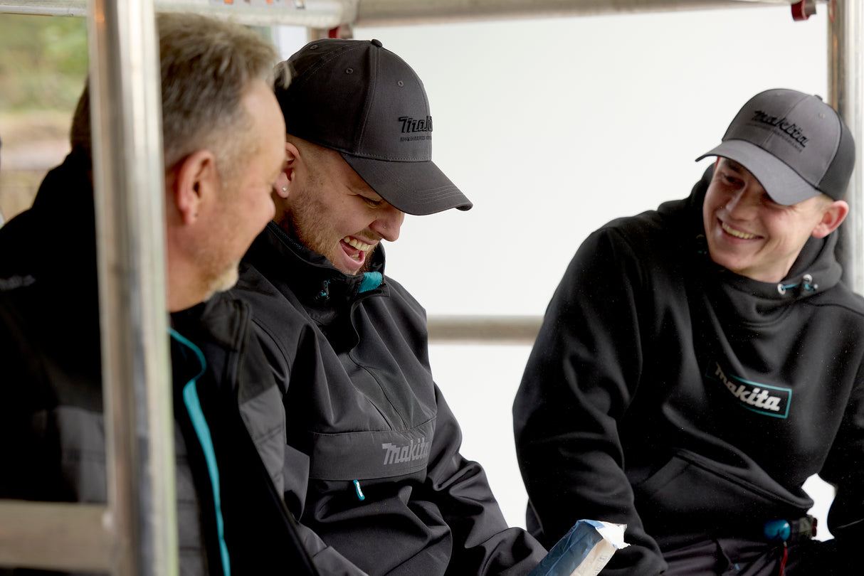 Three men wearing black jackets and Makita hats, sitting together on a vehicle.