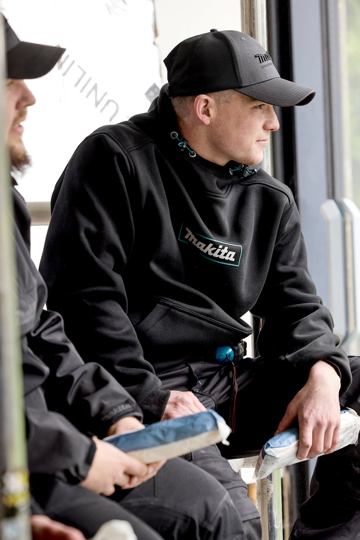 Two men sitting on a bench, one wearing a black hoodie with 'Makita' branding, and a Makita Origin Trucker cap in Grey, also with Makita branding.
