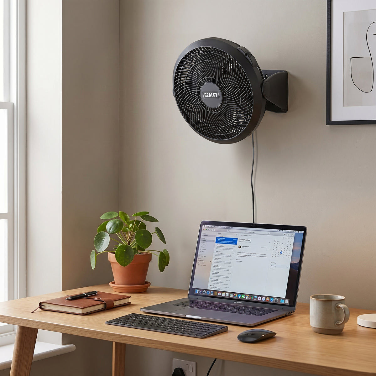Wall-mounted fan above a desk with a laptop, keyboard, and mug.
