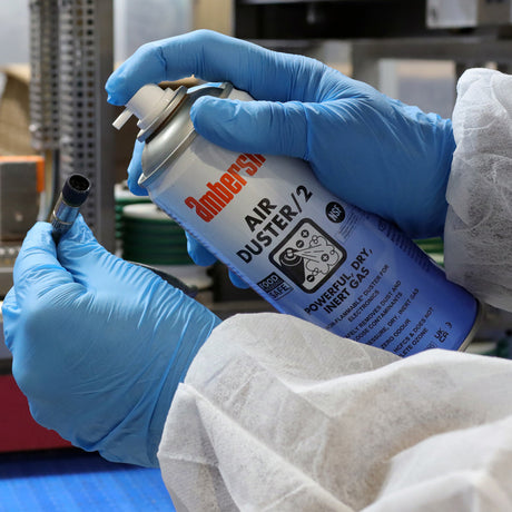 Person in blue gloves and white lab coat using a can of compressed air on a small object in a laboratory setting.