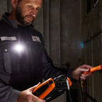 Person wearing a dark jacket with a name tag and logo, holding an orange device in a dimly lit room.