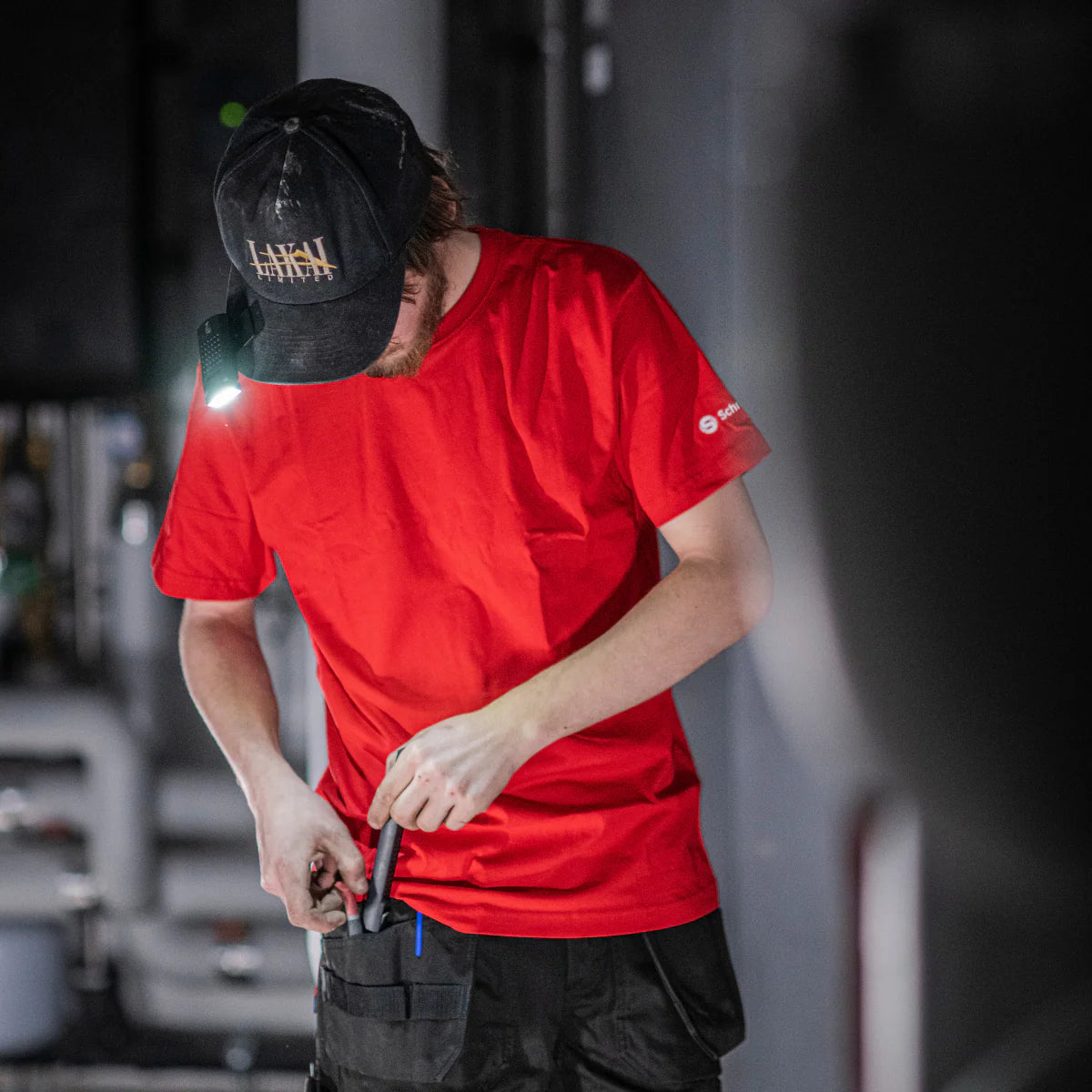 Person wearing a red shirt and black cap with a flashlight, holding a tool in an industrial setting.