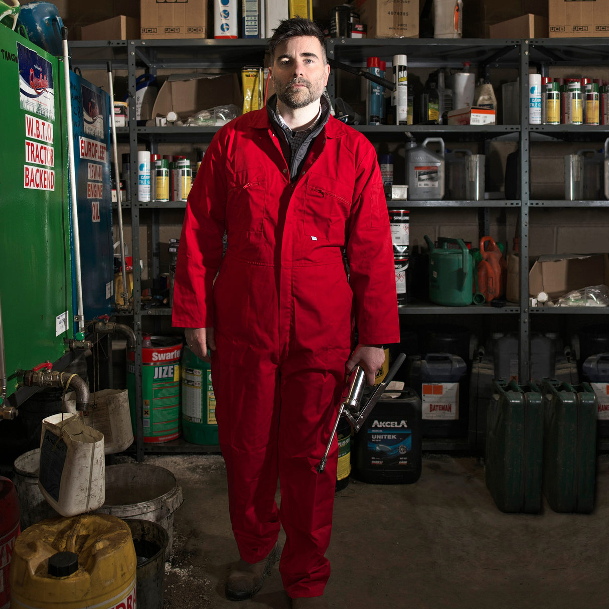 Person wearing a red jumpsuit holding a tool in a workshop setting with shelves and equipment.