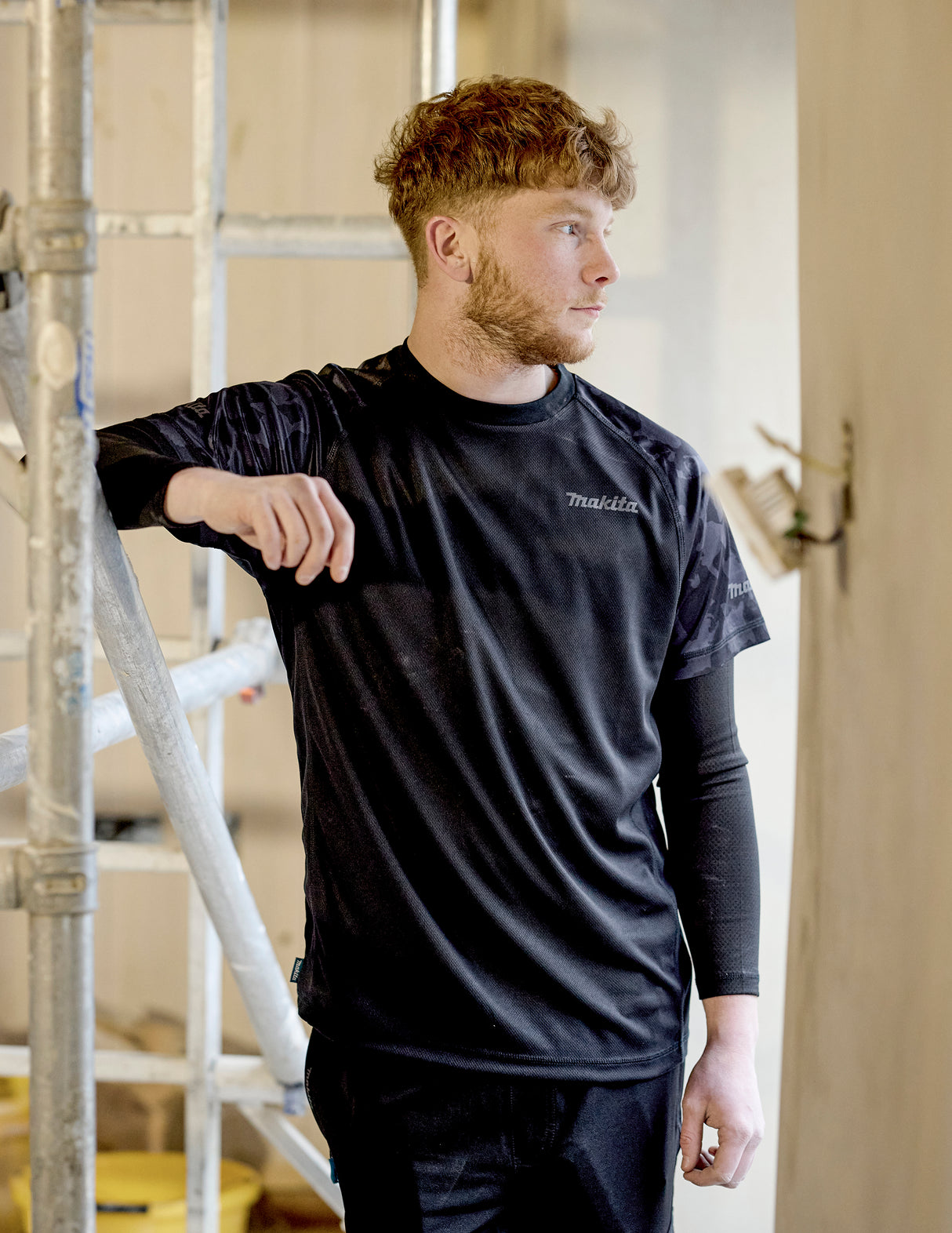 Man wearing a black Makita Tech T-Shirt with a Makita logo, standing indoors near a ladder.