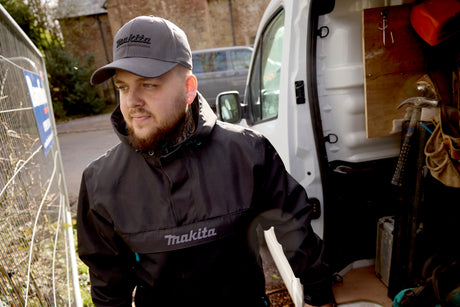 Man wearing a Makita jacket and cap standing next to an open van with tools.