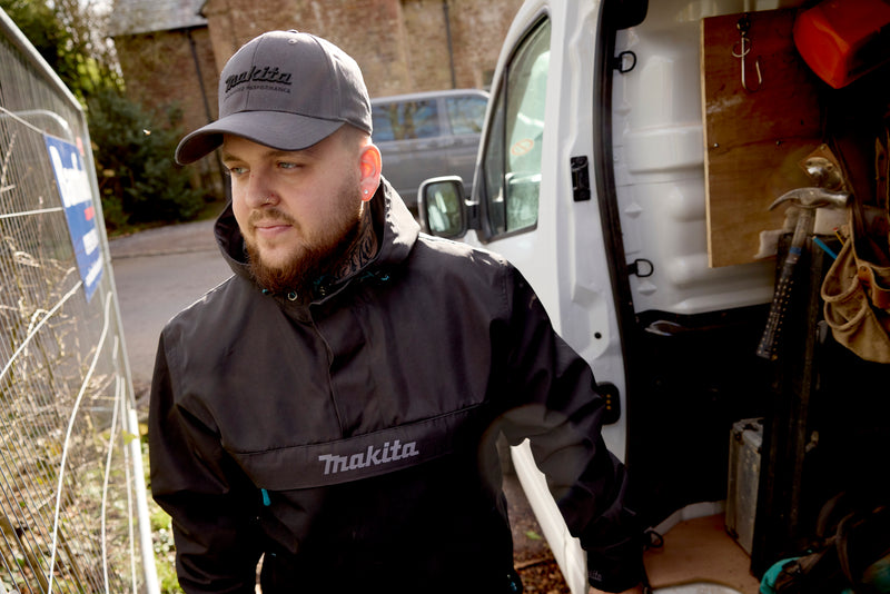 Man wearing a Makita jacket and cap standing next to an open van with tools.