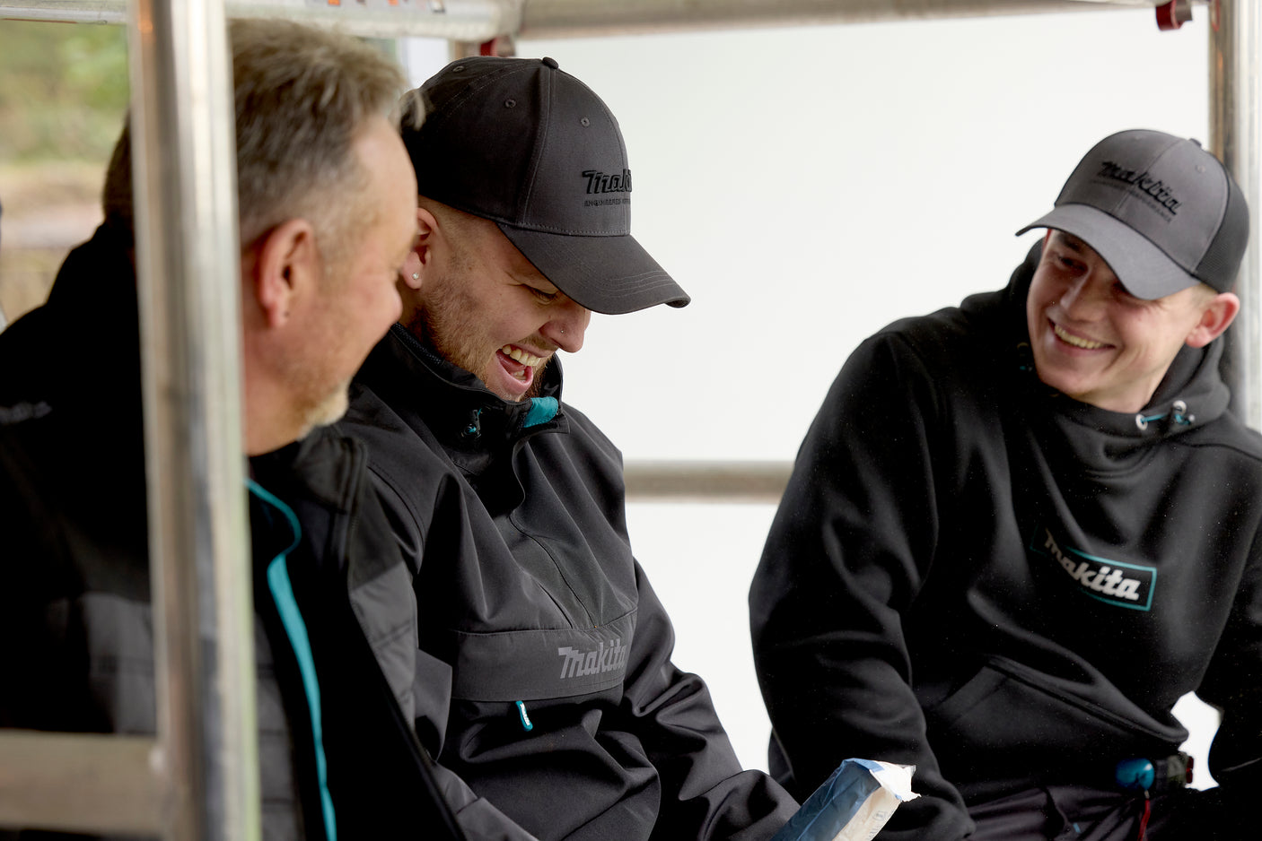 Three men wearing black jackets and Makita hats, sitting together on a vehicle.