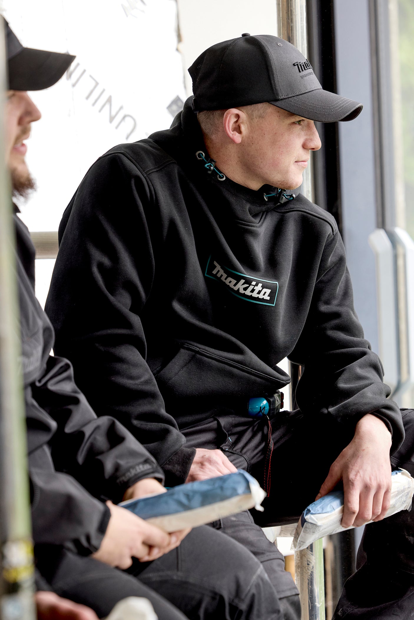 Two men sitting on a bench, one wearing a black hoodie with 'Makita' branding, and a Makita Origin Trucker cap in Grey, also with Makita branding.