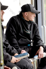 Two men sitting on a bench, one wearing a black hoodie with 'Makita' branding, and a Makita Origin Trucker cap in Grey, also with Makita branding.
