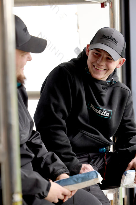 Two individuals wearing black jackets and grey trucker hats with visible Makita branding, sitting on a bench.