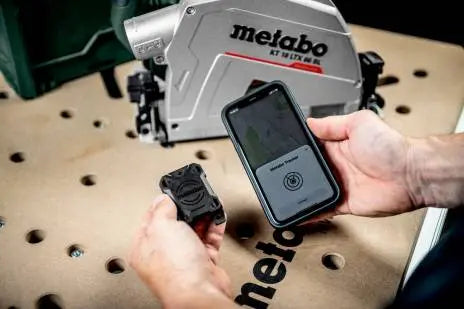 Image showing a pair of hands, in one hand is the Metabo GPS Tracker for ios, in the other is an iphone. The Metabo app is visible on the iPhone screen. A table with a metabo circular saw on it is visable in the background. 