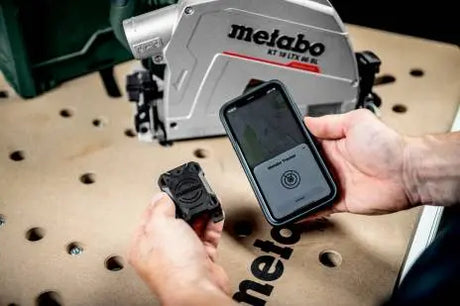 Image showing a pair of hands, in one hand is the Metabo GPS Tracker for ios, in the other is an iphone. The Metabo app is visible on the iPhone screen. A table with a metabo circular saw on it is visable in the background. 