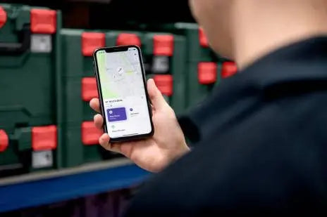 Image showing an iphone being held in a hand. On the screen we can see the Metabo app with tracking information shown. In the background are rows of metabo tools in metaloc boxes. 