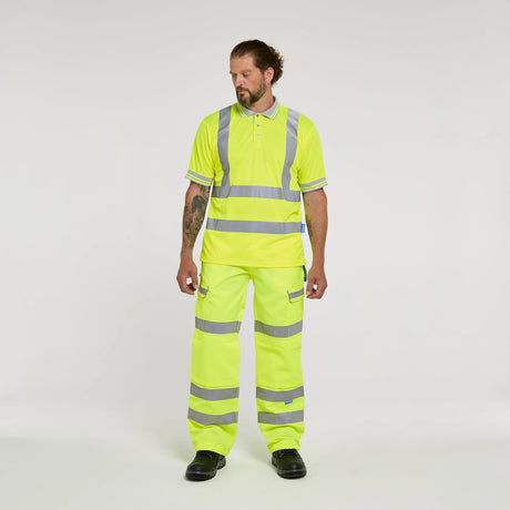 A man modeling a high-visibility neon yellow two-piece work uniform with reflective silver stripes, consisting of a short-sleeve polo shirt and matching trousers, against a plain light gray studio background.