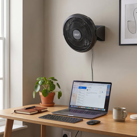 Wall-mounted fan above a desk with a laptop, keyboard, and mug.